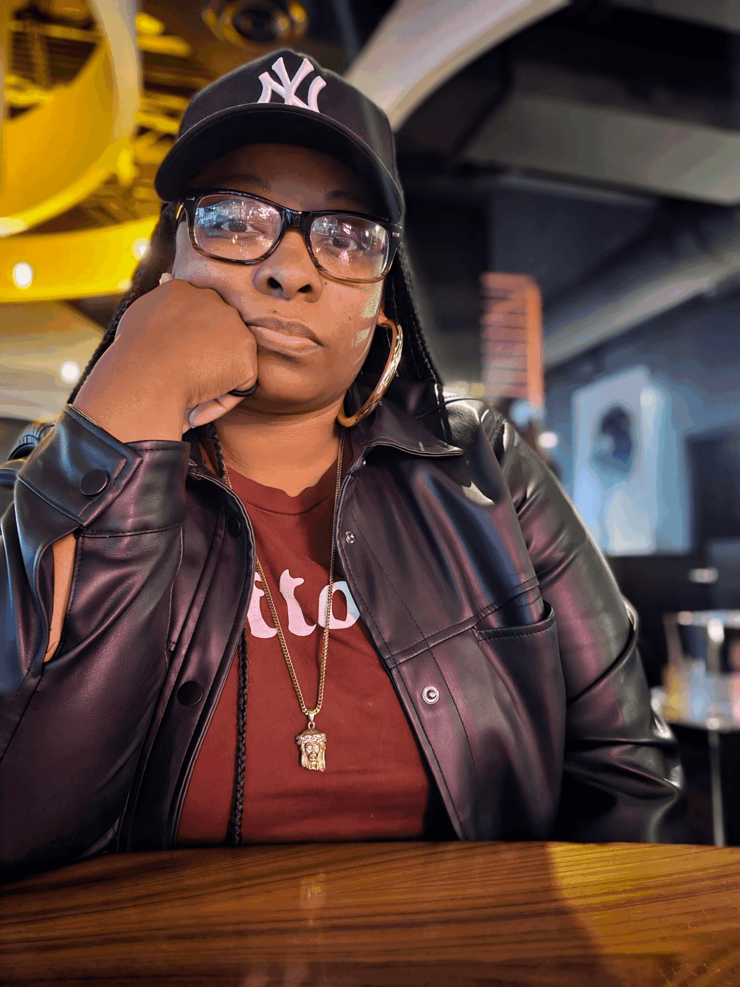 Woman wearing glasses, a black leather jacket, and a New York Yankees cap, sitting at a table in a modern indoor setting.