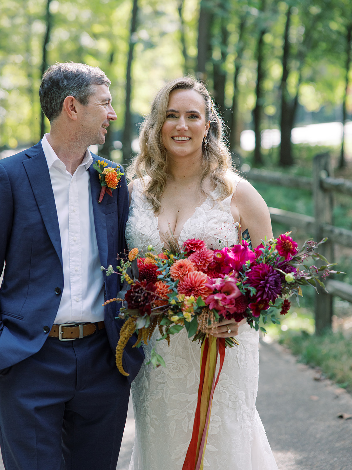 A bride holding a large colorful bouquet stands next to a groom outdoors in a park, smiling, with trees in the background.