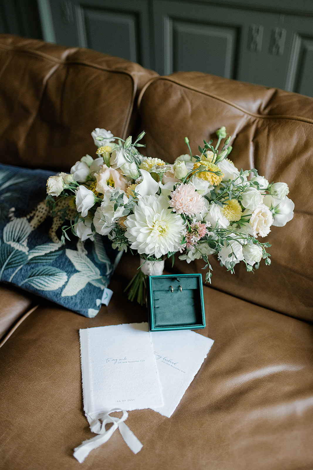 Bouquet of white, pink, and yellow flowers on a brown leather couch with a blue pillow, a jewelry box, and a white cloth.