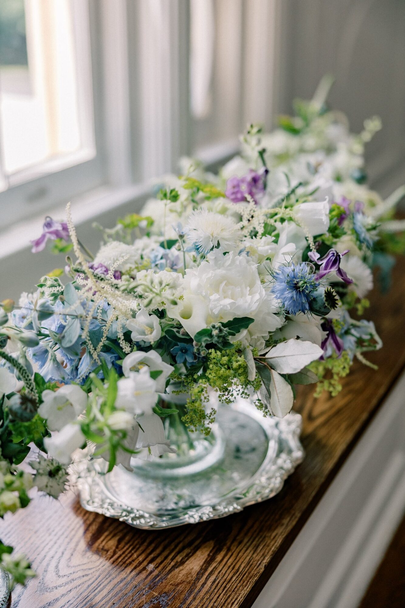 Flower arrangement with white, purple, and blue flowers in a decorative silver bowl on a wooden windowsill