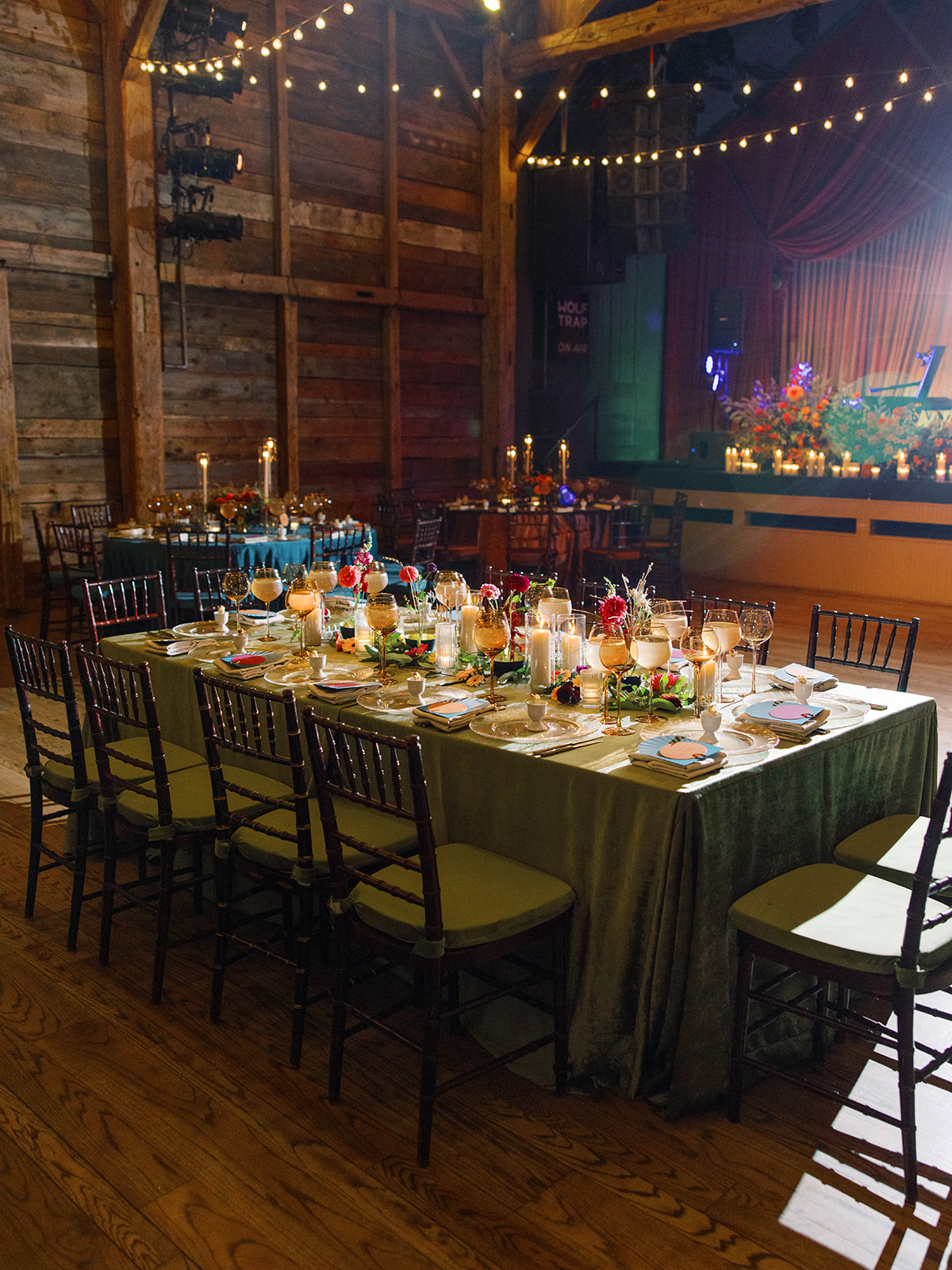Long banquet table set with plates, glasses, and decorations in a rustic venue with string lights and a stage in the background.