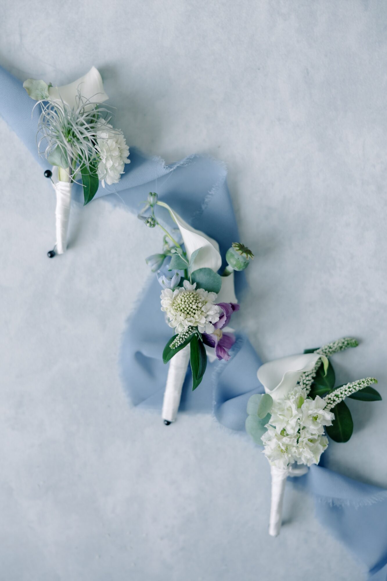 Three floral boutonnieres with white and purple flowers on light blue fabric, on a light gray surface.