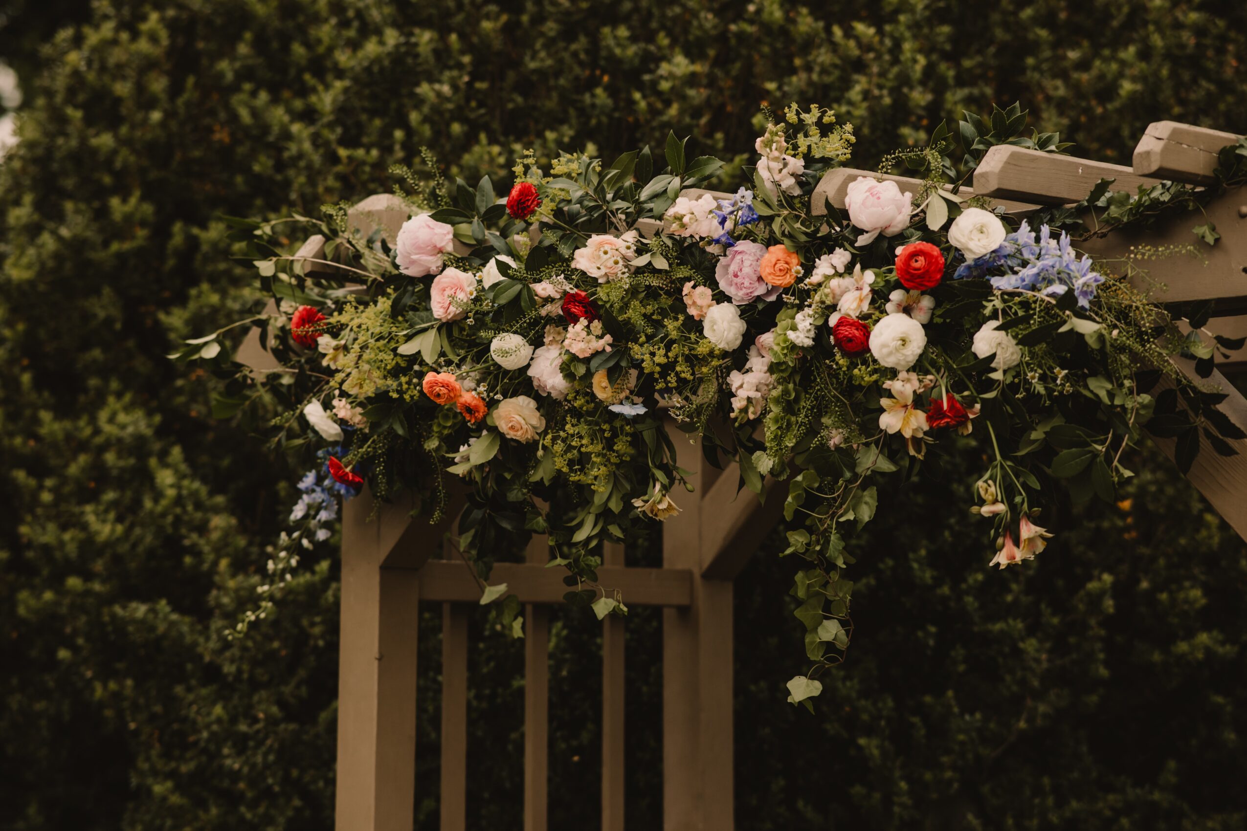 Two large floral arrangements with pink, white, red, and purple flowers on a wooden arch outdoors, green trees in background.