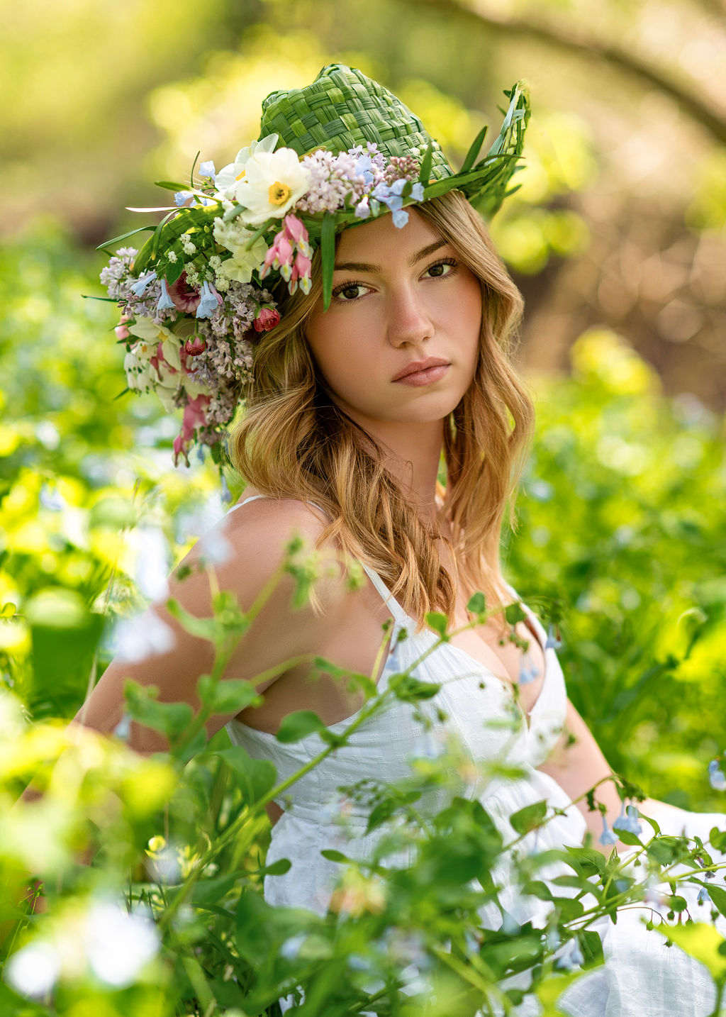 Young woman with long hair wearing a floral hat and white dress outdoors surrounded by greenery.