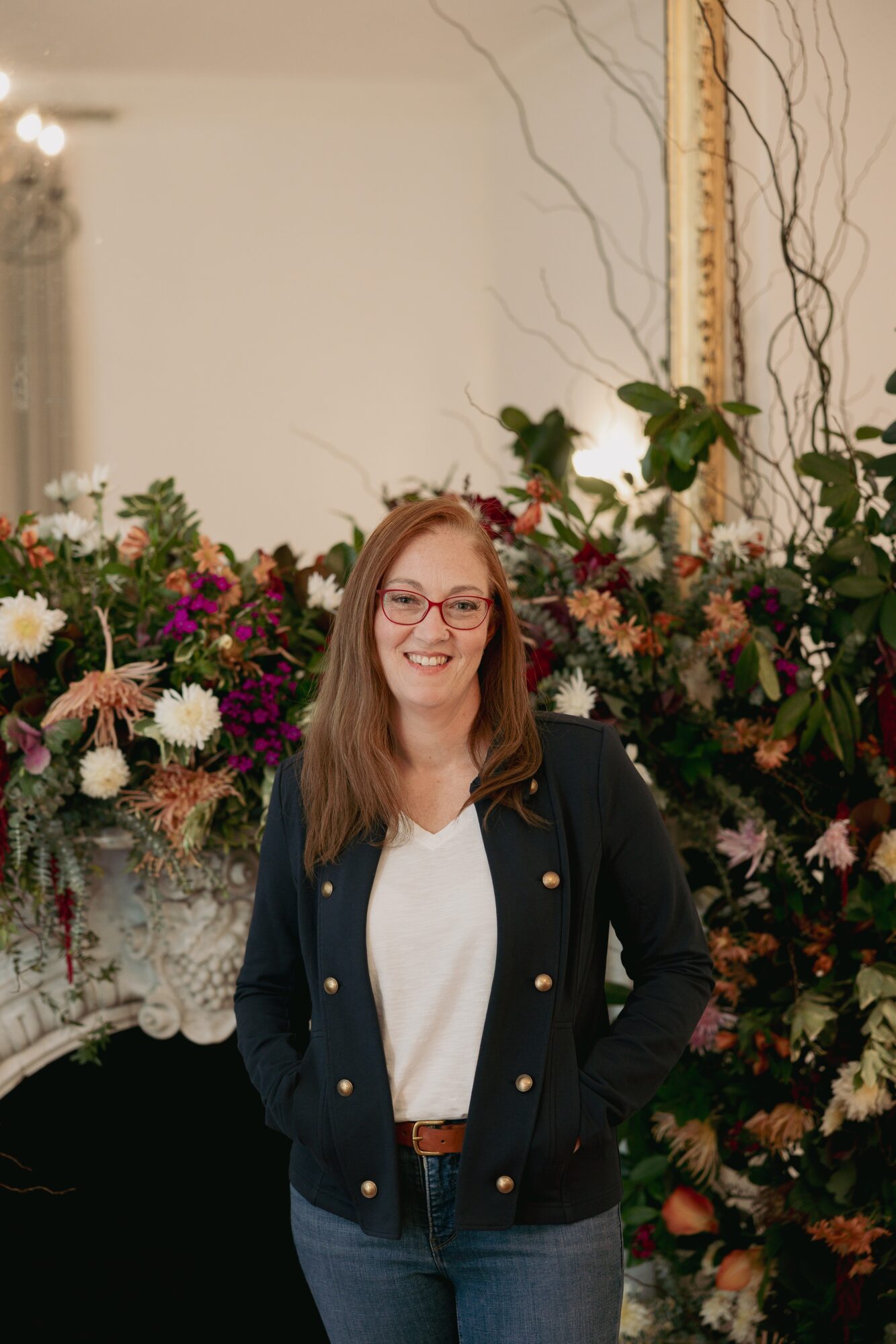 Woman with glasses smiling, standing in front of a large floral arrangement indoors.