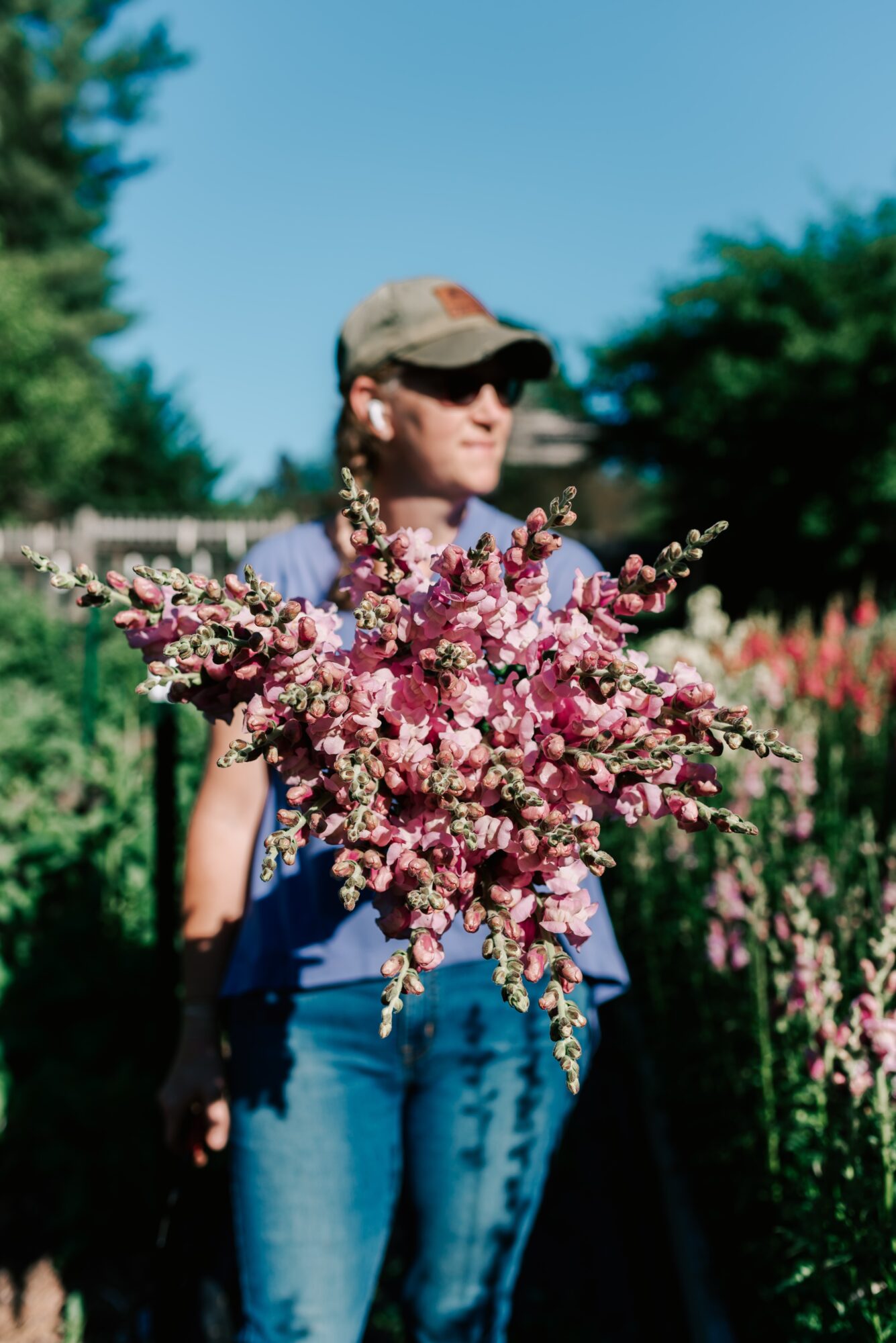 Woman wearing sunglasses, a cap, and earrings holding a large pink flower bouquet outdoors on a sunny day.