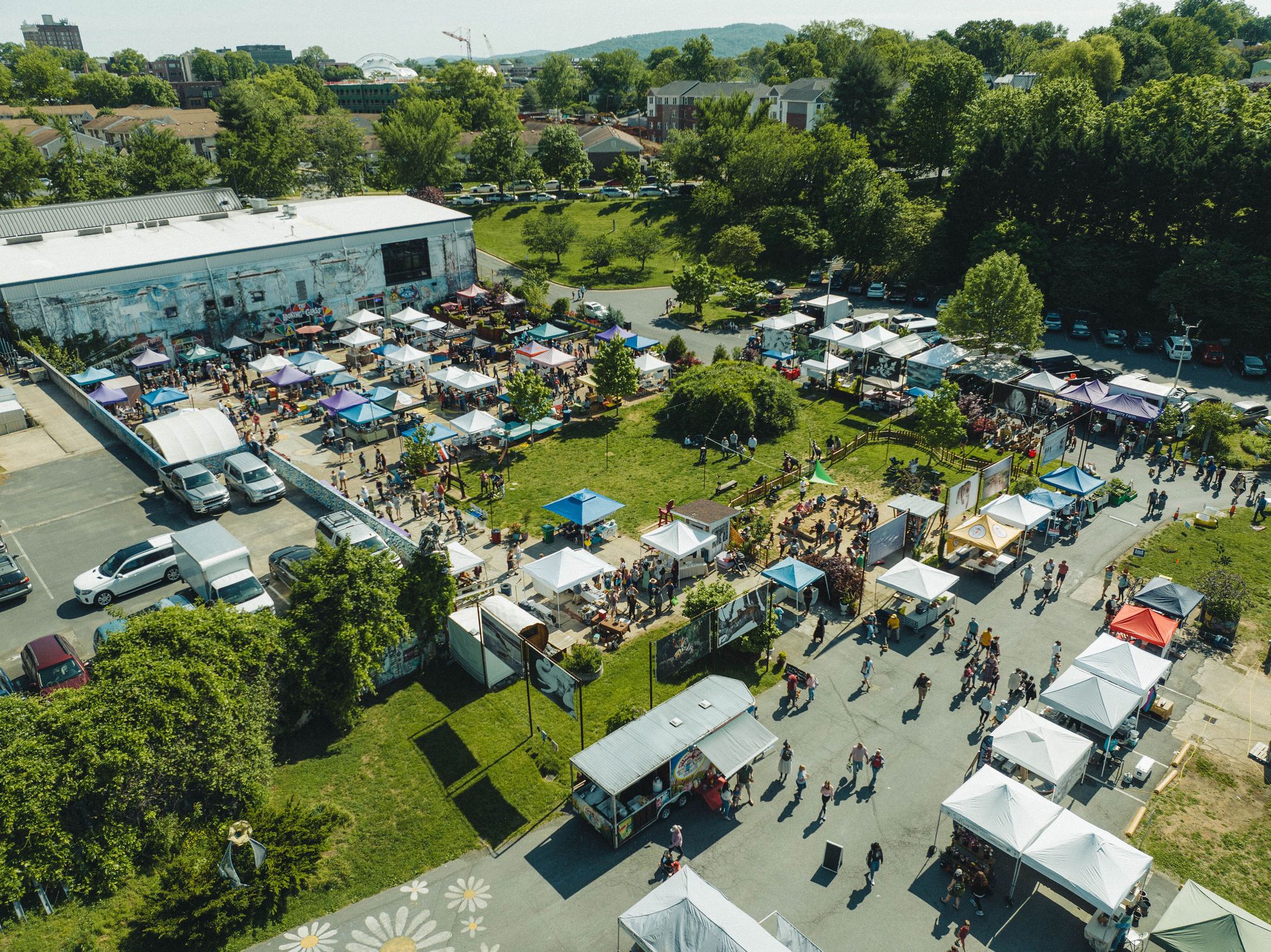 Outdoor market with tents, people walking, and a large building in the background, surrounded by trees and greenery.