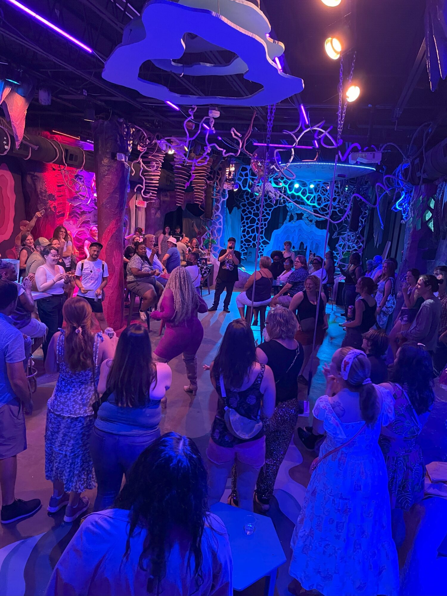 Crowd of people dancing and socializing in a colorful, illuminated indoor space with decorative hanging elements.