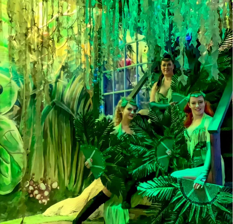 Four women in green costumes pose among jungle-themed decorations with plants and vines.