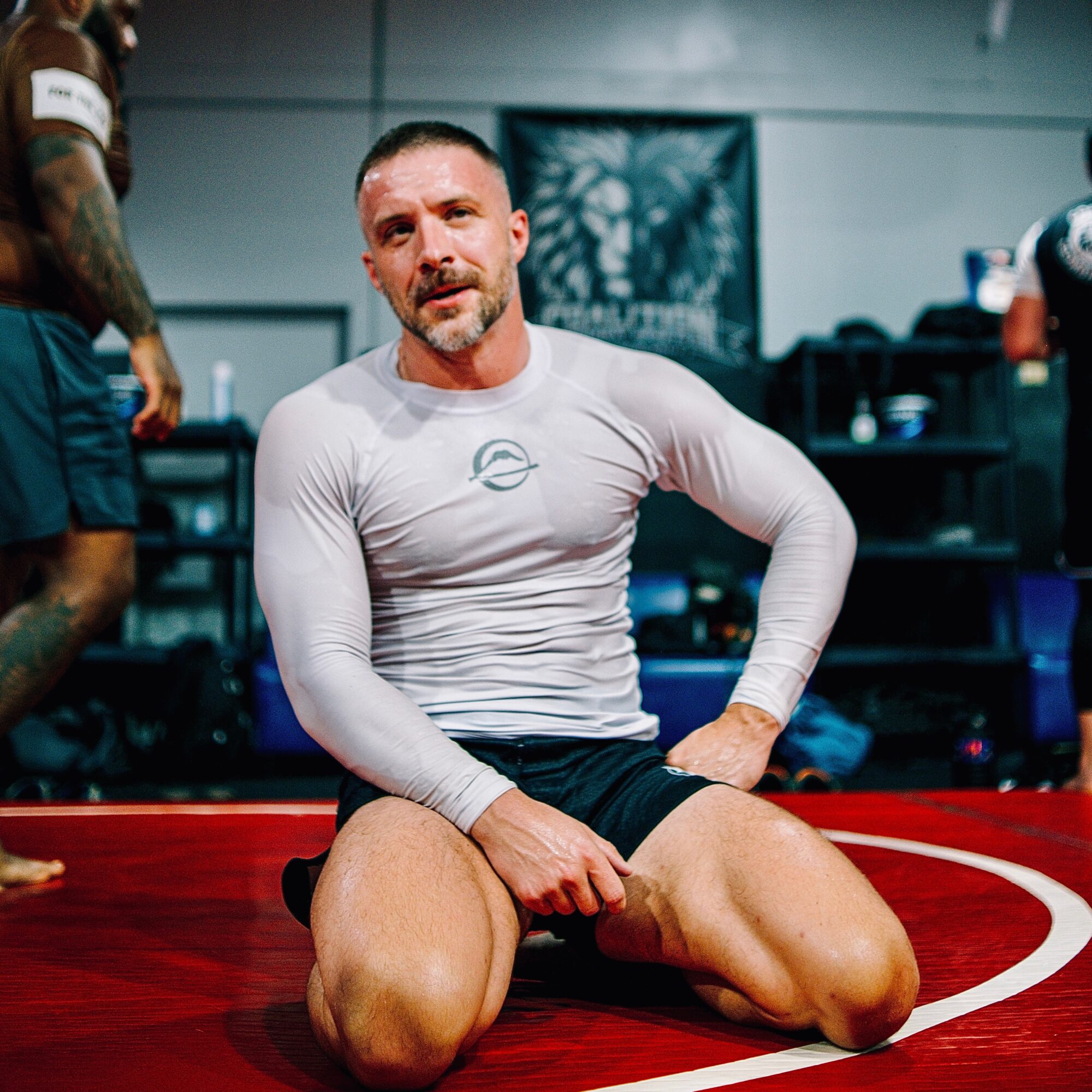 Man kneeling on a red sports mat in a gym, wearing a white long-sleeve shirt and black shorts, with a serious expression.