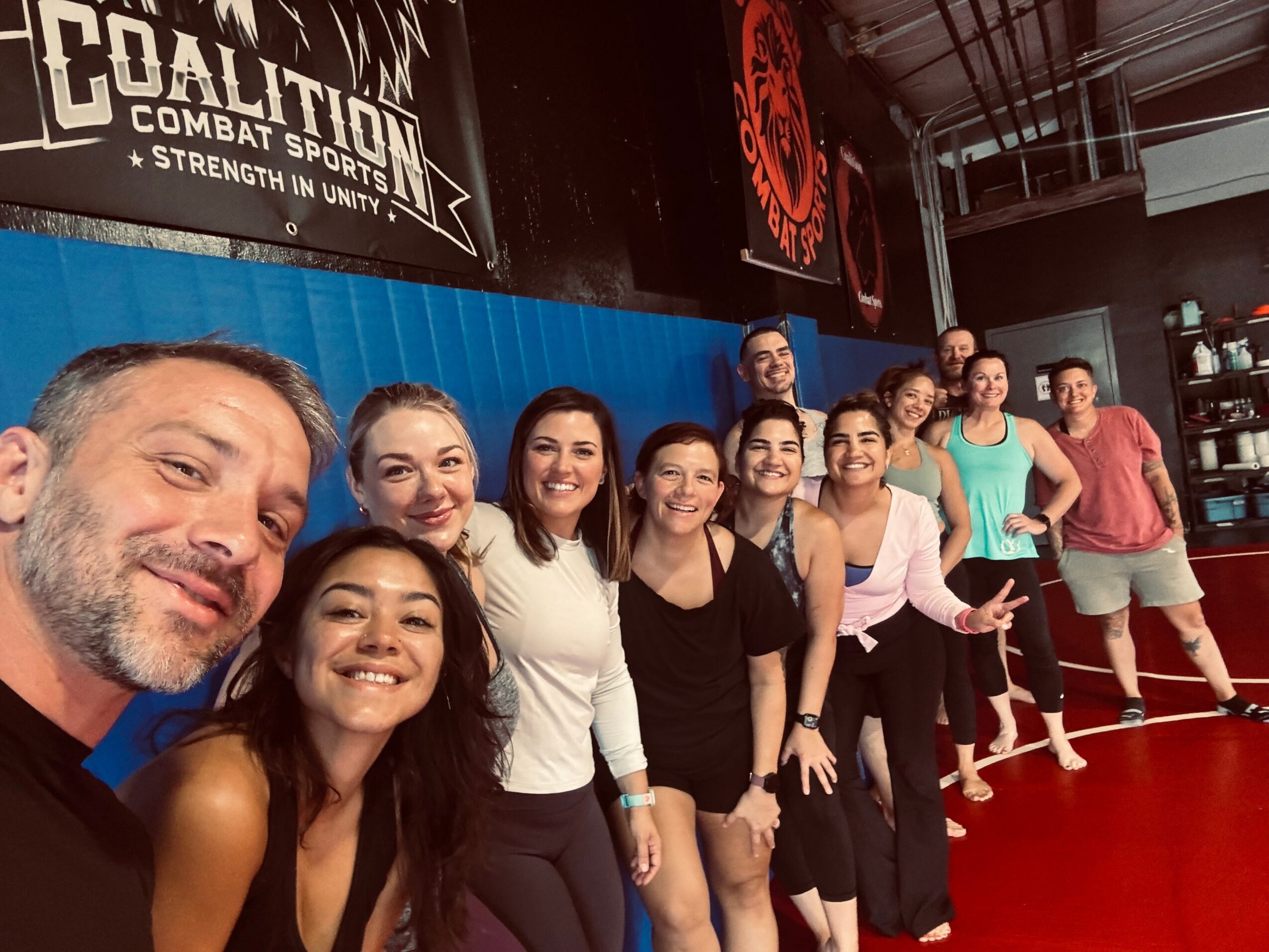 Group of people smiling and posing together indoors, some making peace signs, in a gym or martial arts studio.