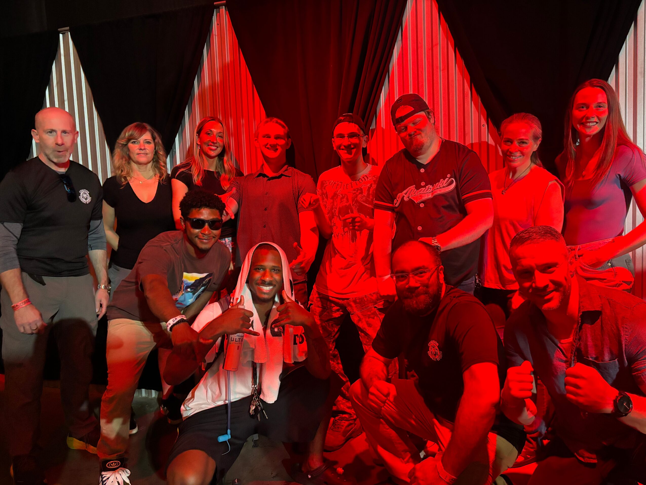 Group of ten people on stage with red and black curtains, some smiling and posing, some wearing sunglasses.