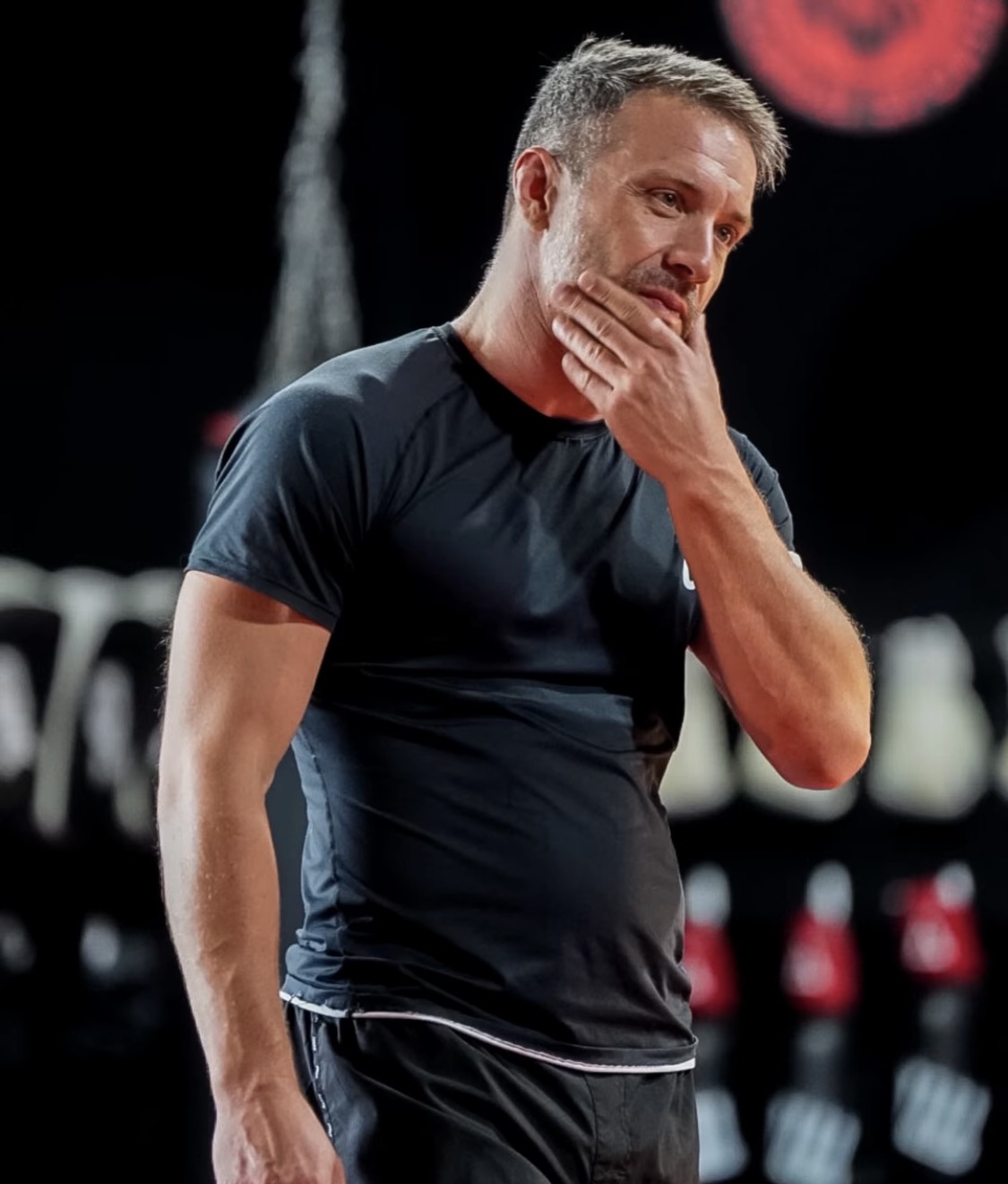 Man with short gray hair and beard, wearing a black shirt, touching his chin thoughtfully, in a gym or fitness setting.