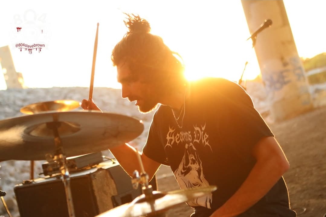 Person playing drums outdoors with sunlight behind, wearing a black T-shirt with a graphic design.