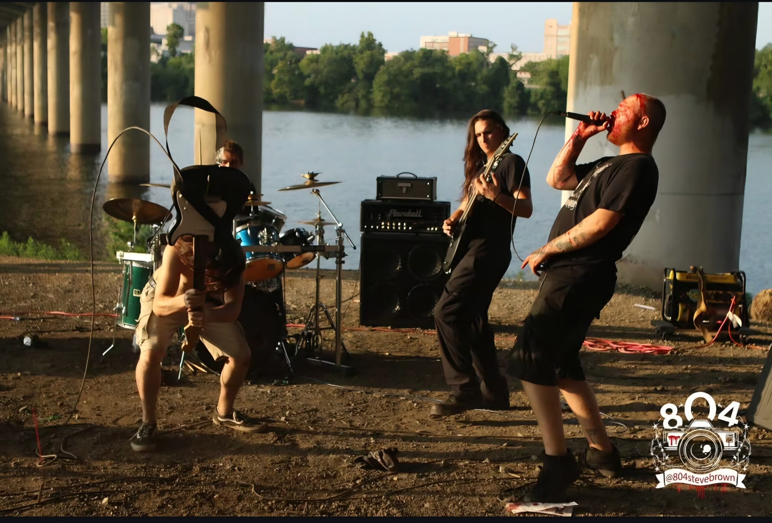 Band performs outdoors near water with trees and buildings in background, under large concrete pillars, during daytime.
