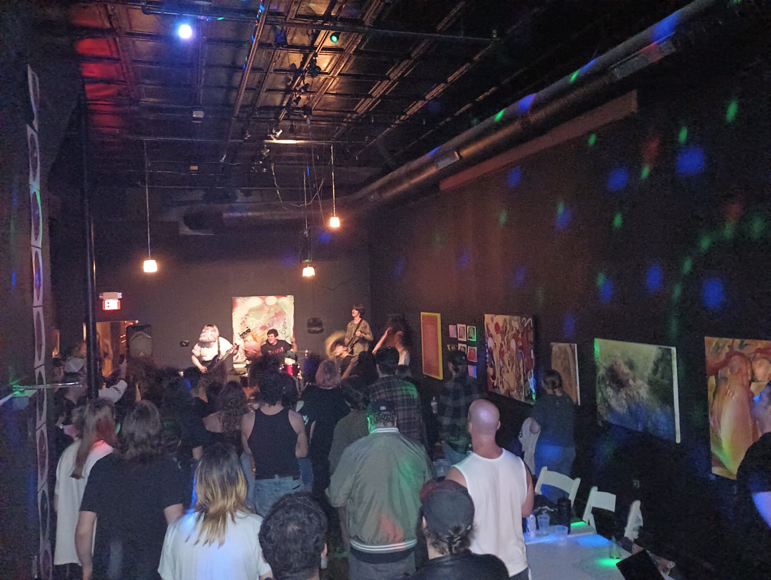Crowd gathered in a dark room with colorful artwork on walls, people watching a performance on stage, some with drinks, dim lighting.
