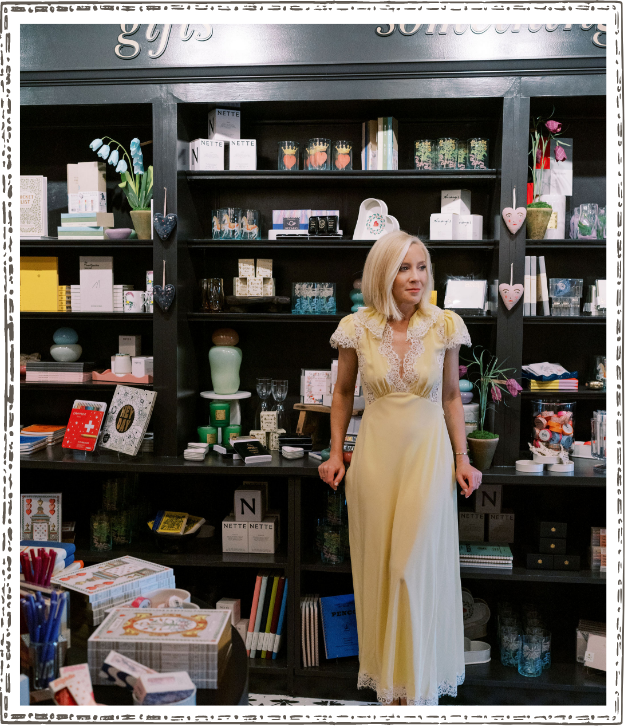 Woman with blonde hair in a yellow dress standing in front of black shelves filled with various items.