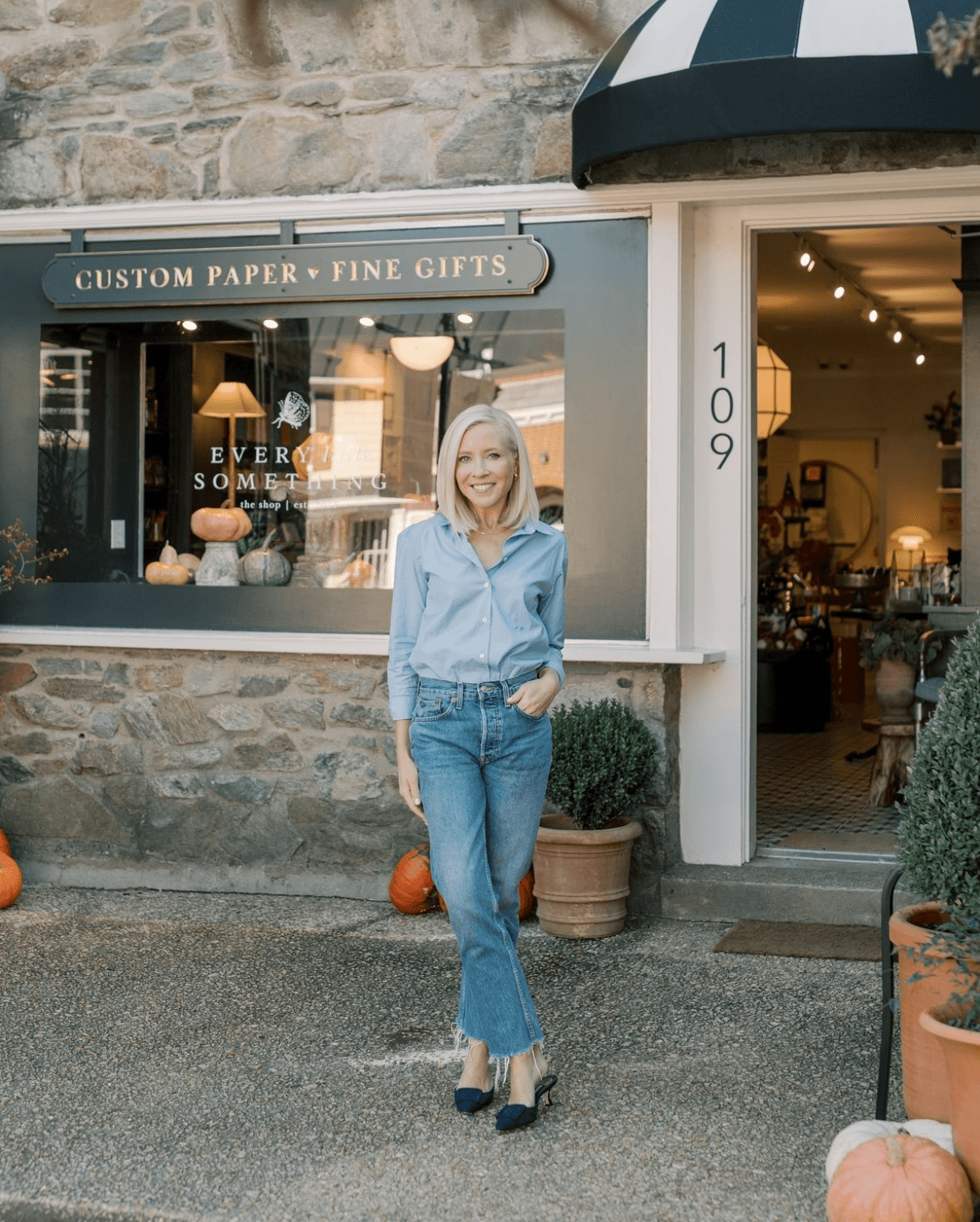 Woman with gray hair in a blue shirt and jeans standing outside a store with stone walls and potted plants.