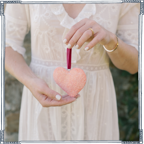 Person holding a heart-shaped object with a pink stick, wearing a white lace dress, outdoors.
