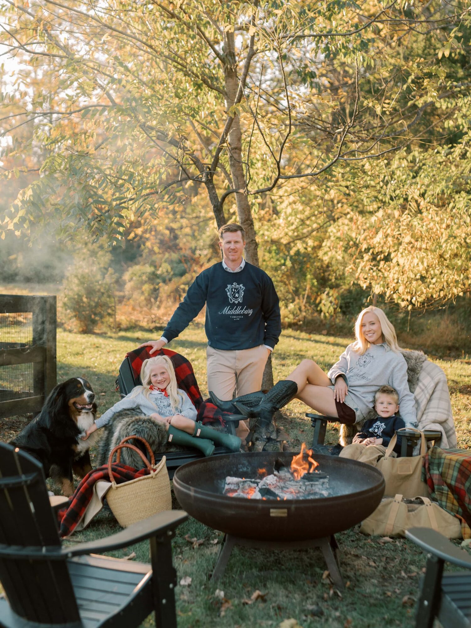 Four people and a dog gather outdoors around a fire pit in a park with trees and autumn leaves, smiling and relaxing.