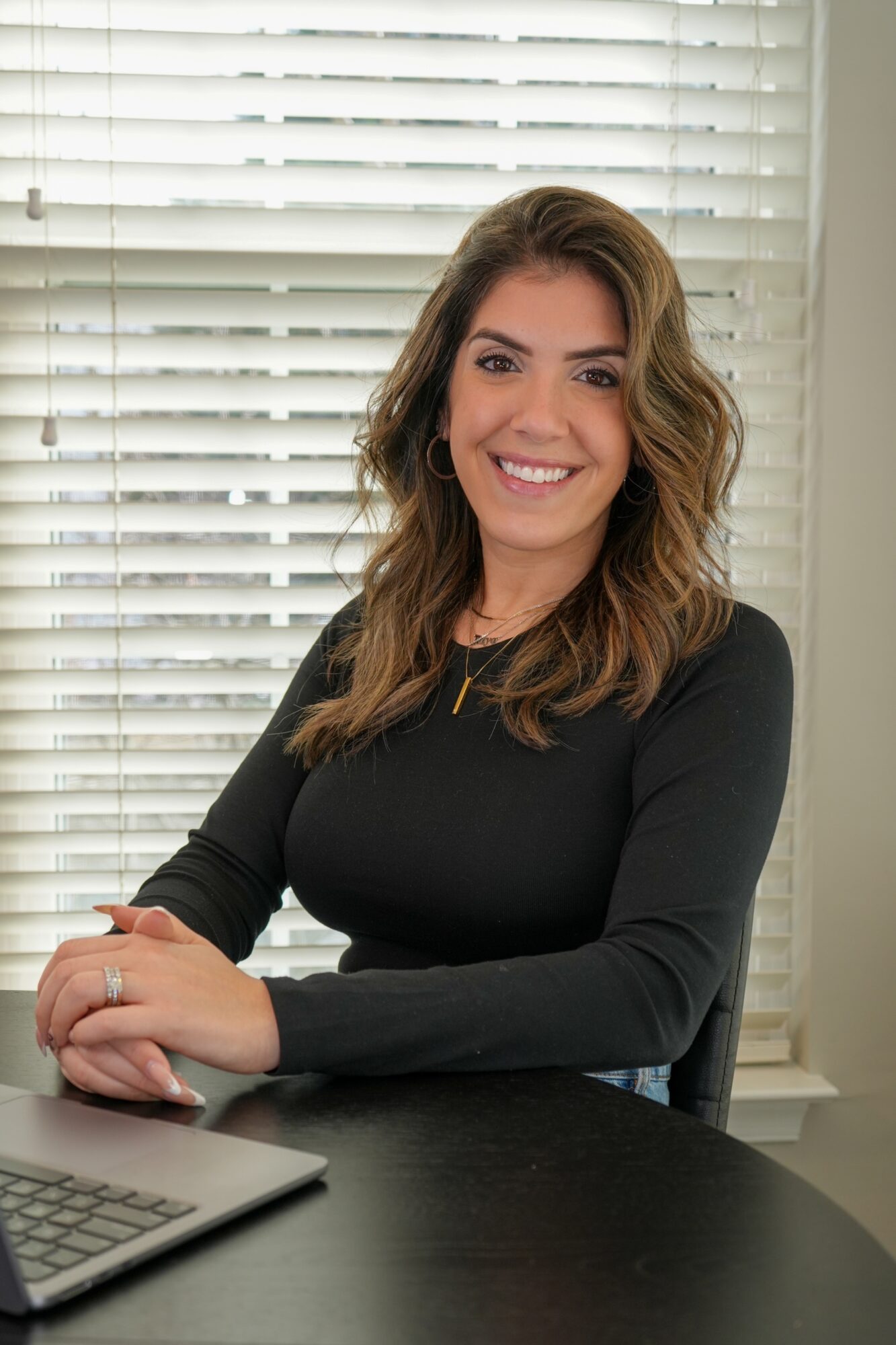 Woman with wavy hair smiling, sitting at a desk with blinds in the background.