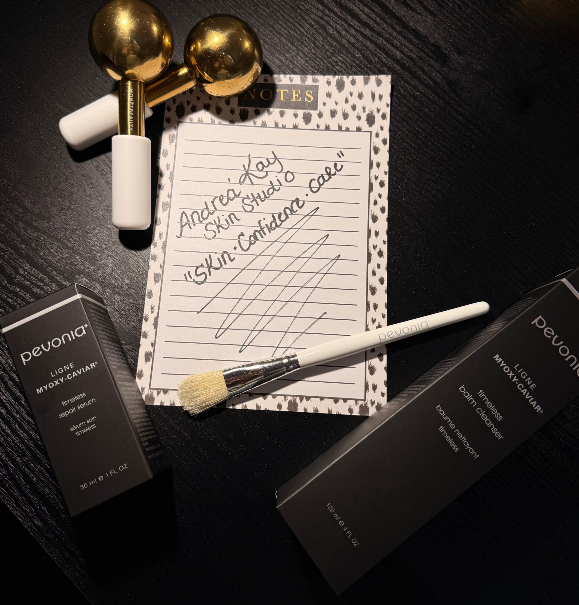 Skincare products, a note, and a makeup brush on a dark surface. Note has handwritten text and lines. Two gold and white tools are nearby.
