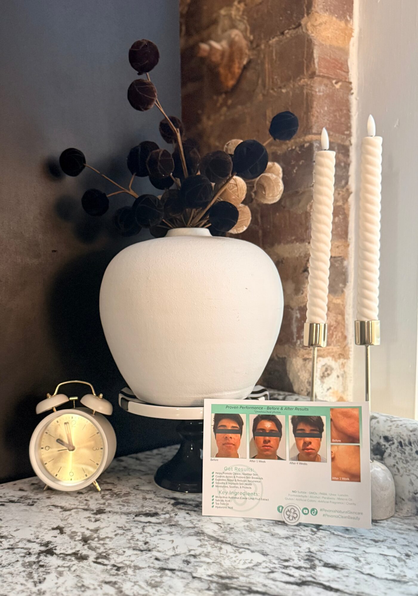 Decorative arrangement with a white vase holding dark dried flowers, two white candles in holders, a small clock, and a photo card, against a brick wall.