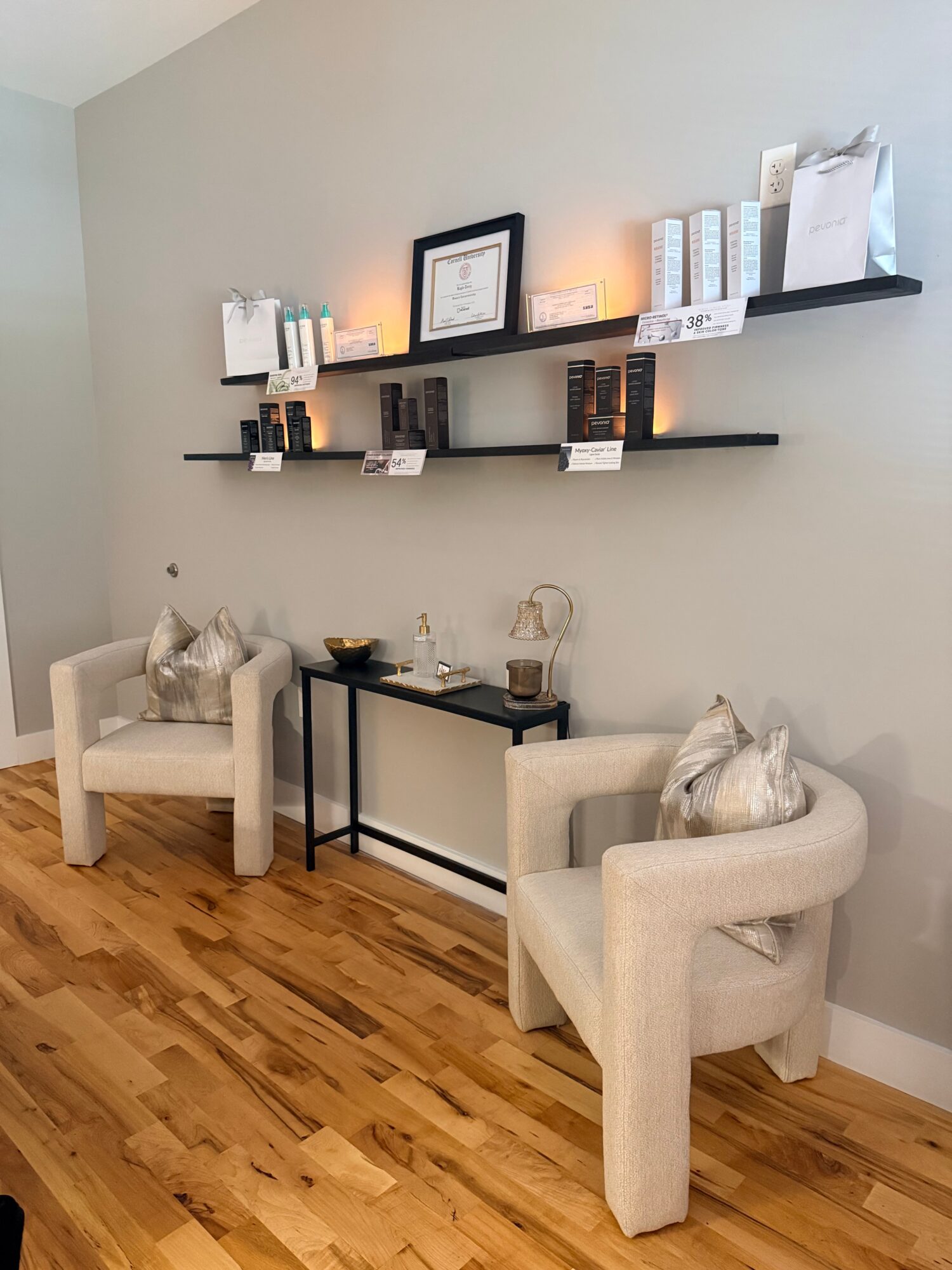 Two beige armchairs with pillows, a black table with decorative items, and two black shelves with books and framed items on a light gray wall.