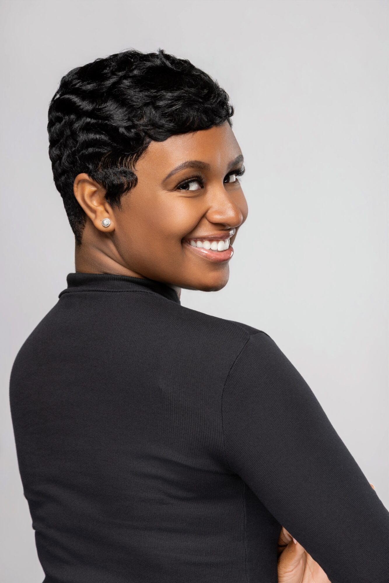 Smiling woman with short, curly hair wearing a black top, facing slightly to the right, against a plain light background.
