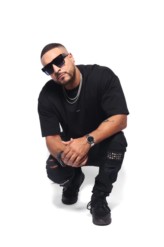 Man crouching, wearing sunglasses, black clothing, and jewelry, against a white background.