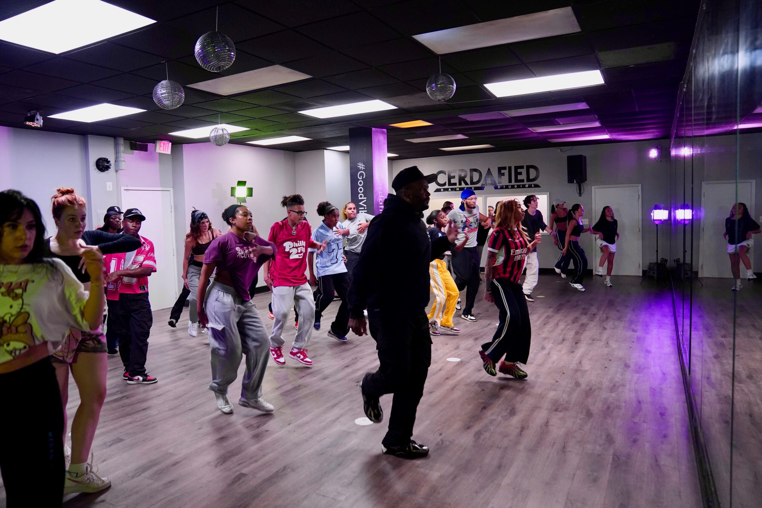 People dancing in a dance studio with purple and black lighting, mirrors, and a wooden floor.