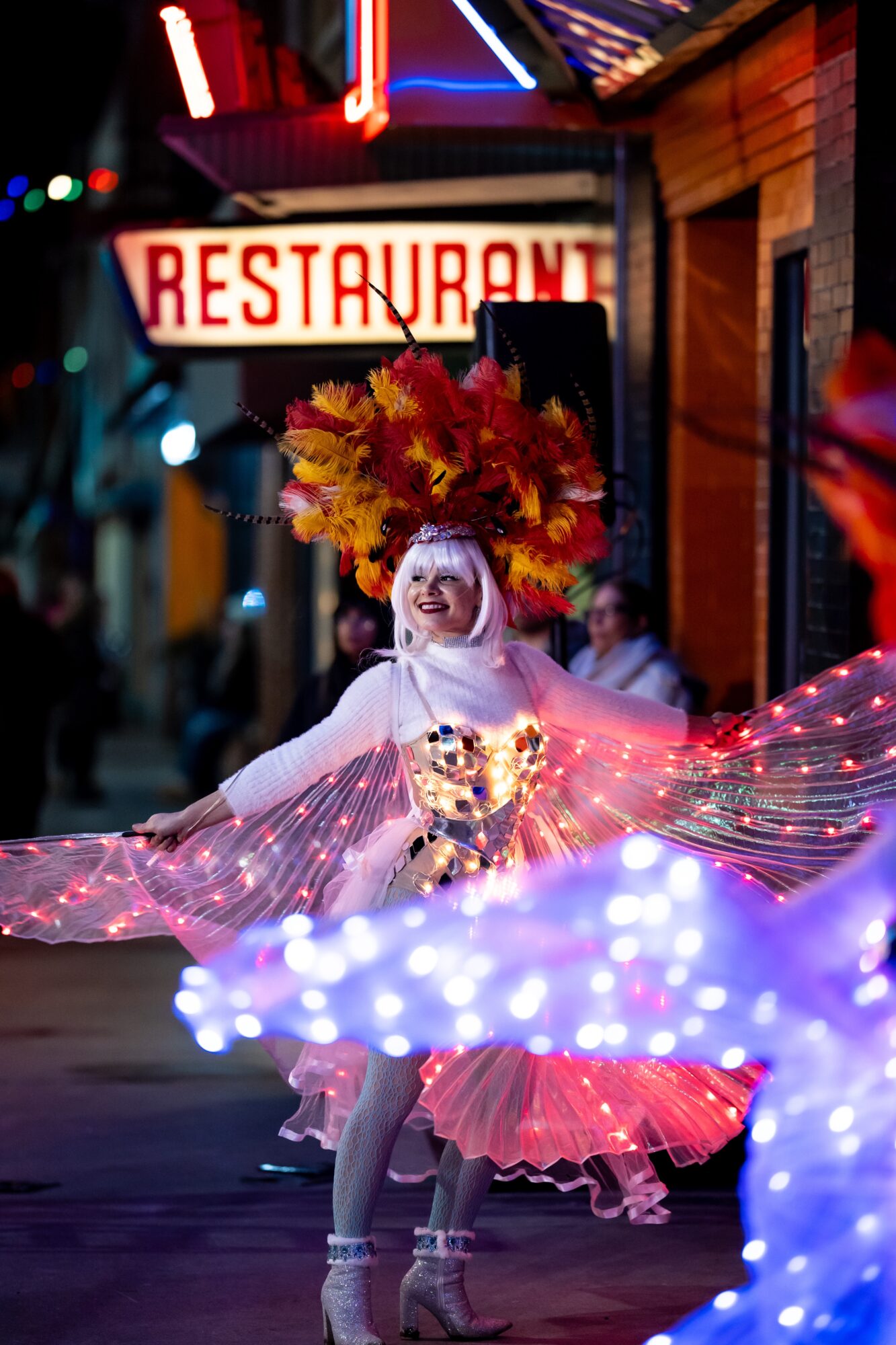 Performer in colorful costume with large feathered headdress dancing on street at night, illuminated by lights.