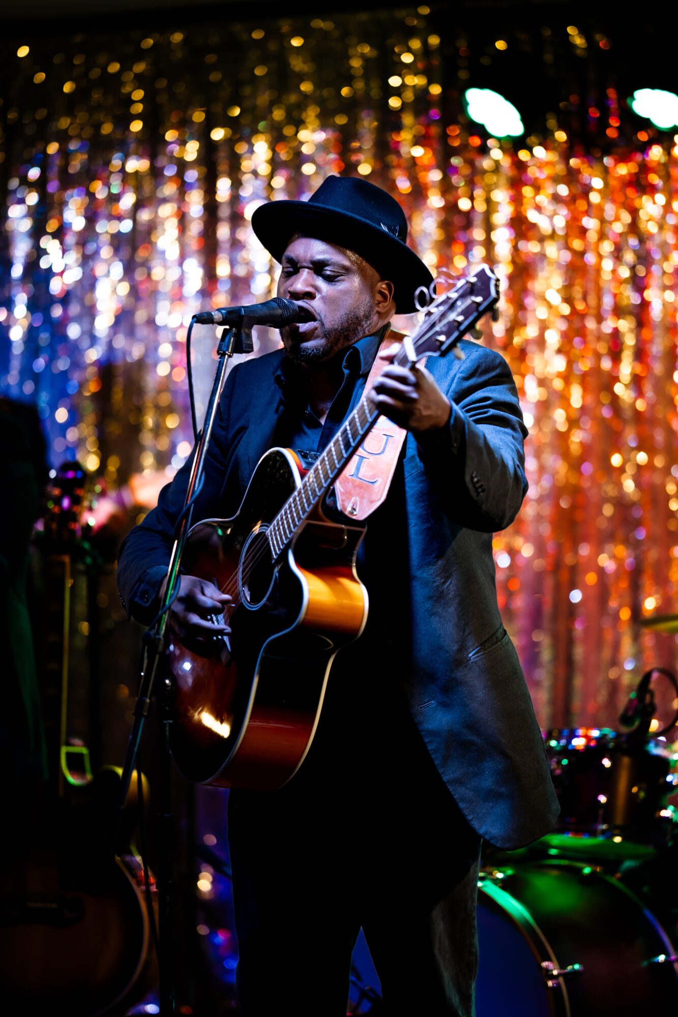 Man wearing a hat and suit playing guitar and singing into a microphone on stage with colorful background lights.