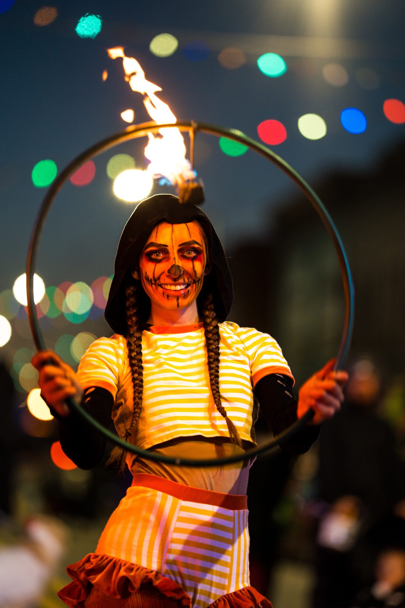 Circus performer with face paint holding a fire hoop, colorful lights in background, smiling, wearing striped shirt and skirt.