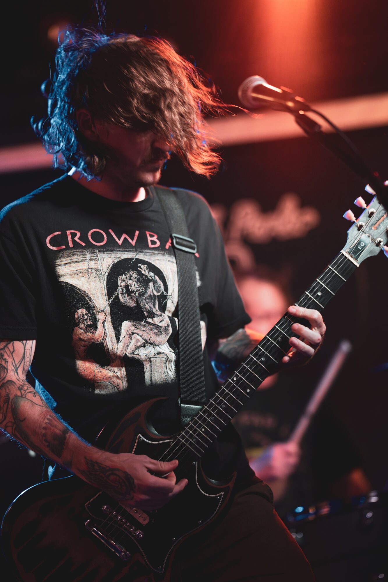 Person playing electric guitar on stage with red and blue lighting, wearing a black T-shirt with skull design.
