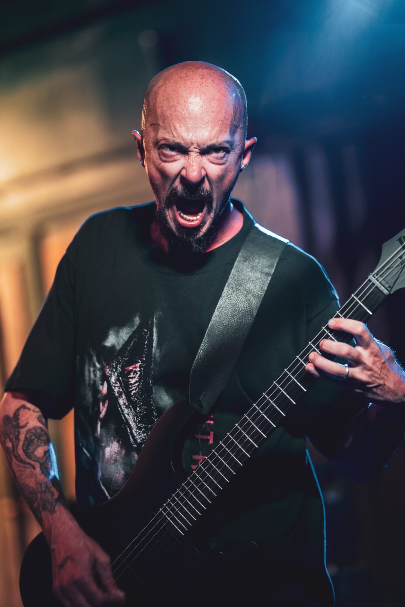 Bald man with beard passionately playing guitar on stage, mouth open as if shouting or singing, dark background with stage lighting
