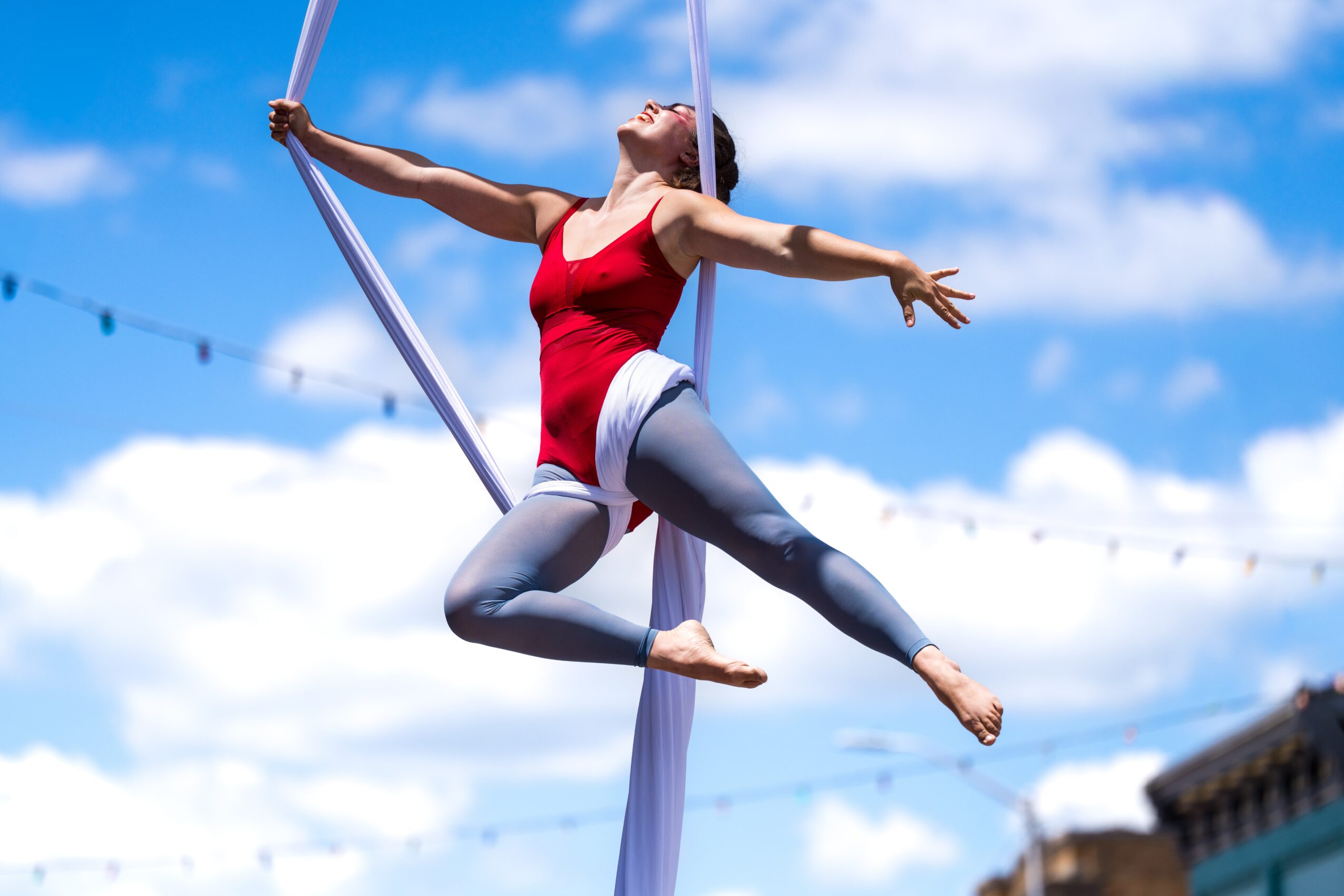 A woman performs aerial silk acrobatics outdoors against a blue sky with clouds, wearing a red top and gray leggings.