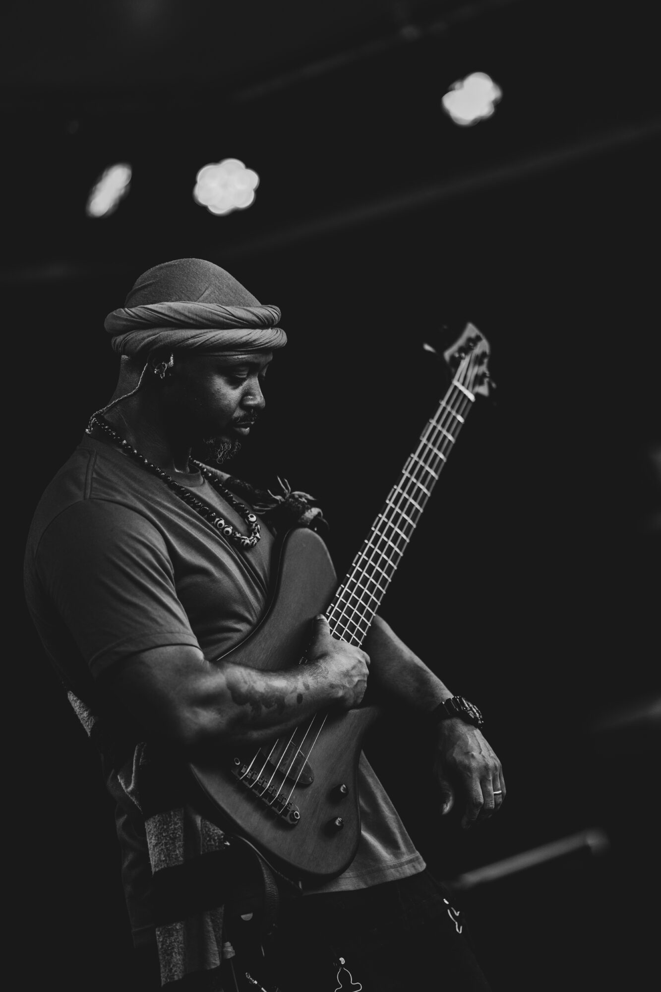 Person playing an electric guitar on stage, wearing a headwrap and jewelry, with stage lights overhead.