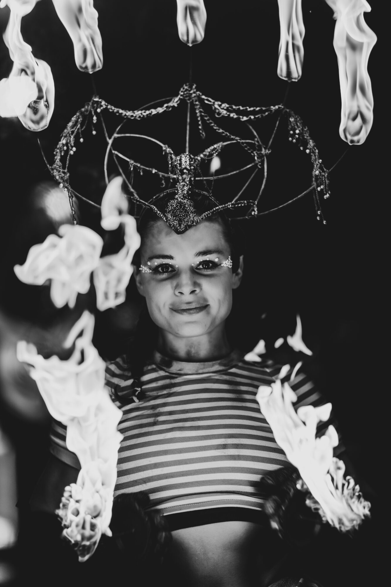 Child with a headpiece made of chains and hanging objects, wearing a striped shirt, surrounded by floating objects, in black and white.