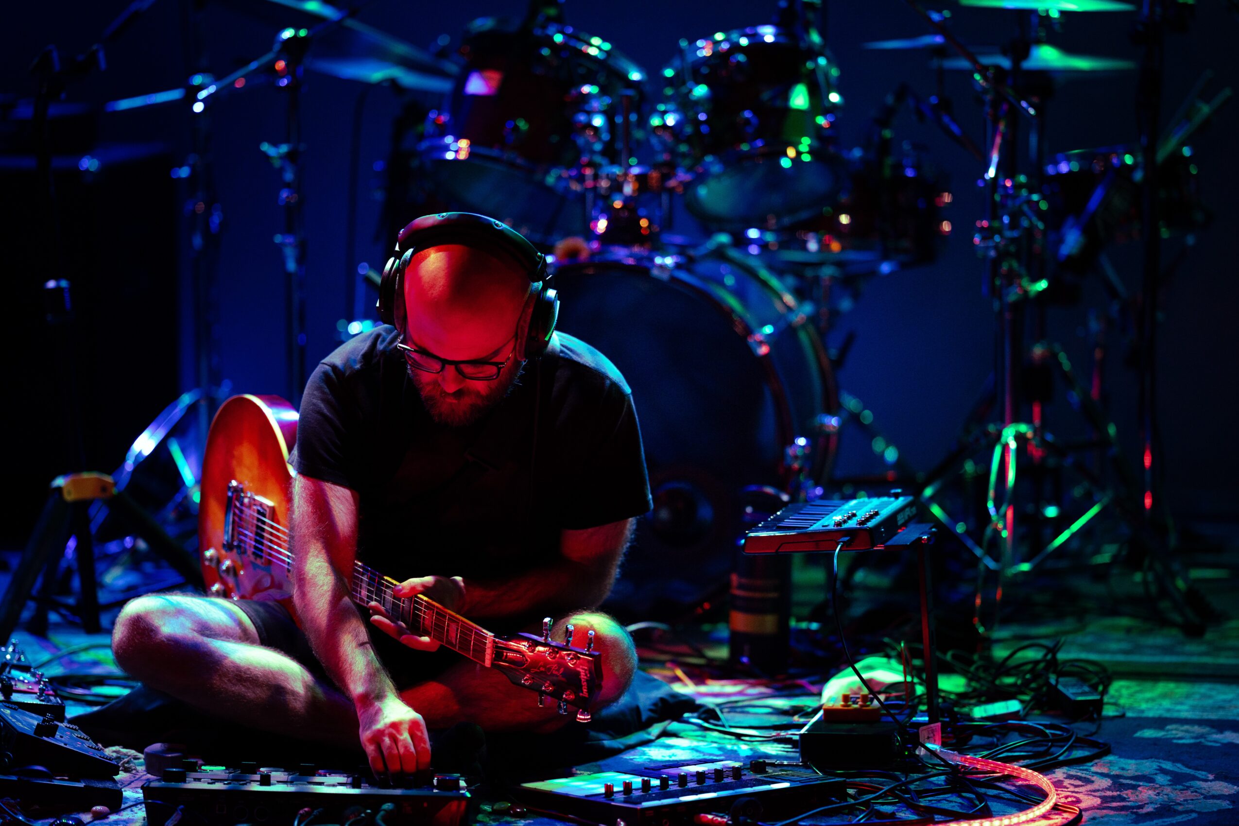 Person sitting cross-legged on stage with guitar, surrounded by musical equipment and drums, under colorful stage lighting.