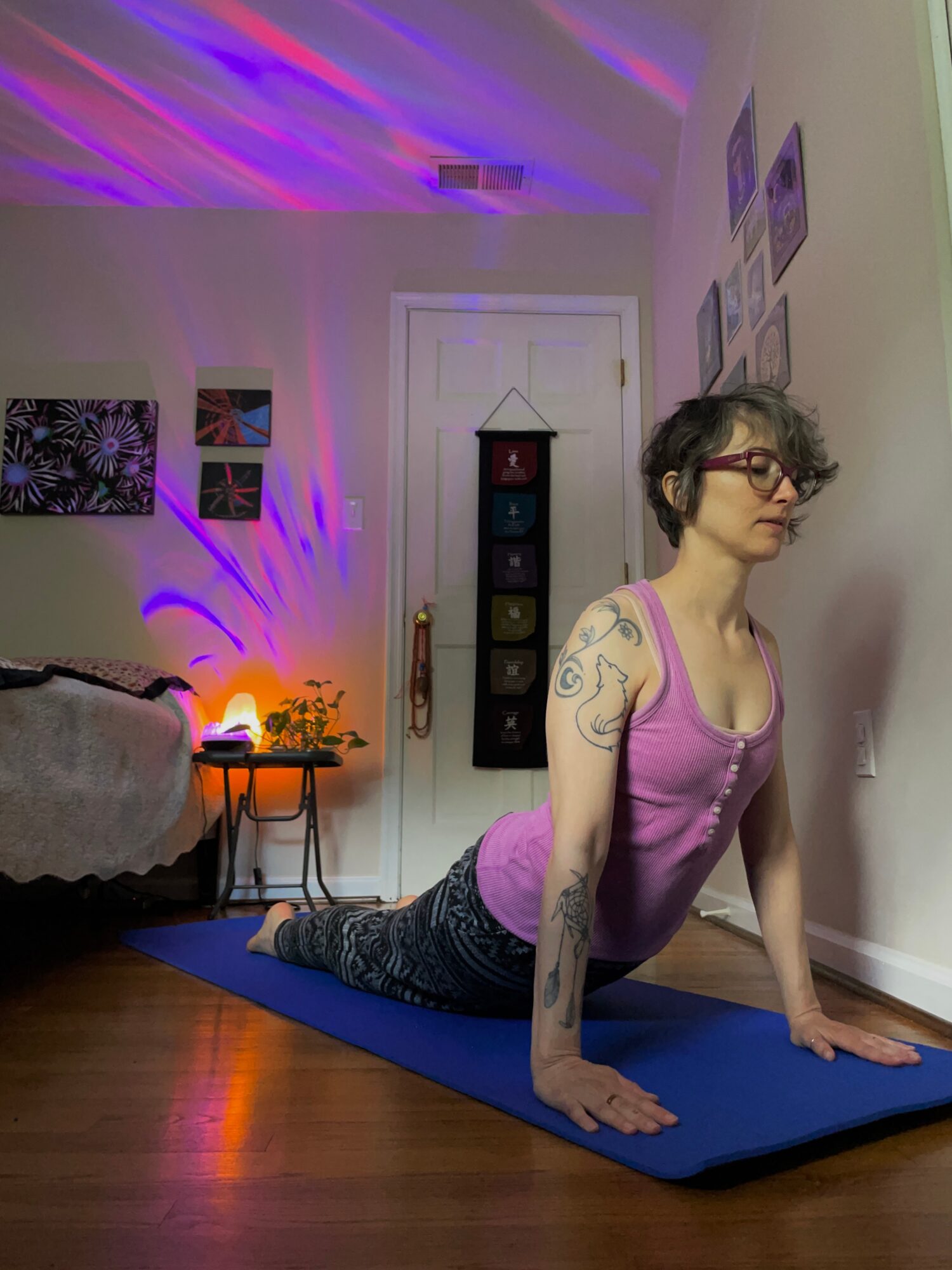 Woman practicing yoga on a mat in a living room with wall art and a shelf, illuminated by colorful lighting.
