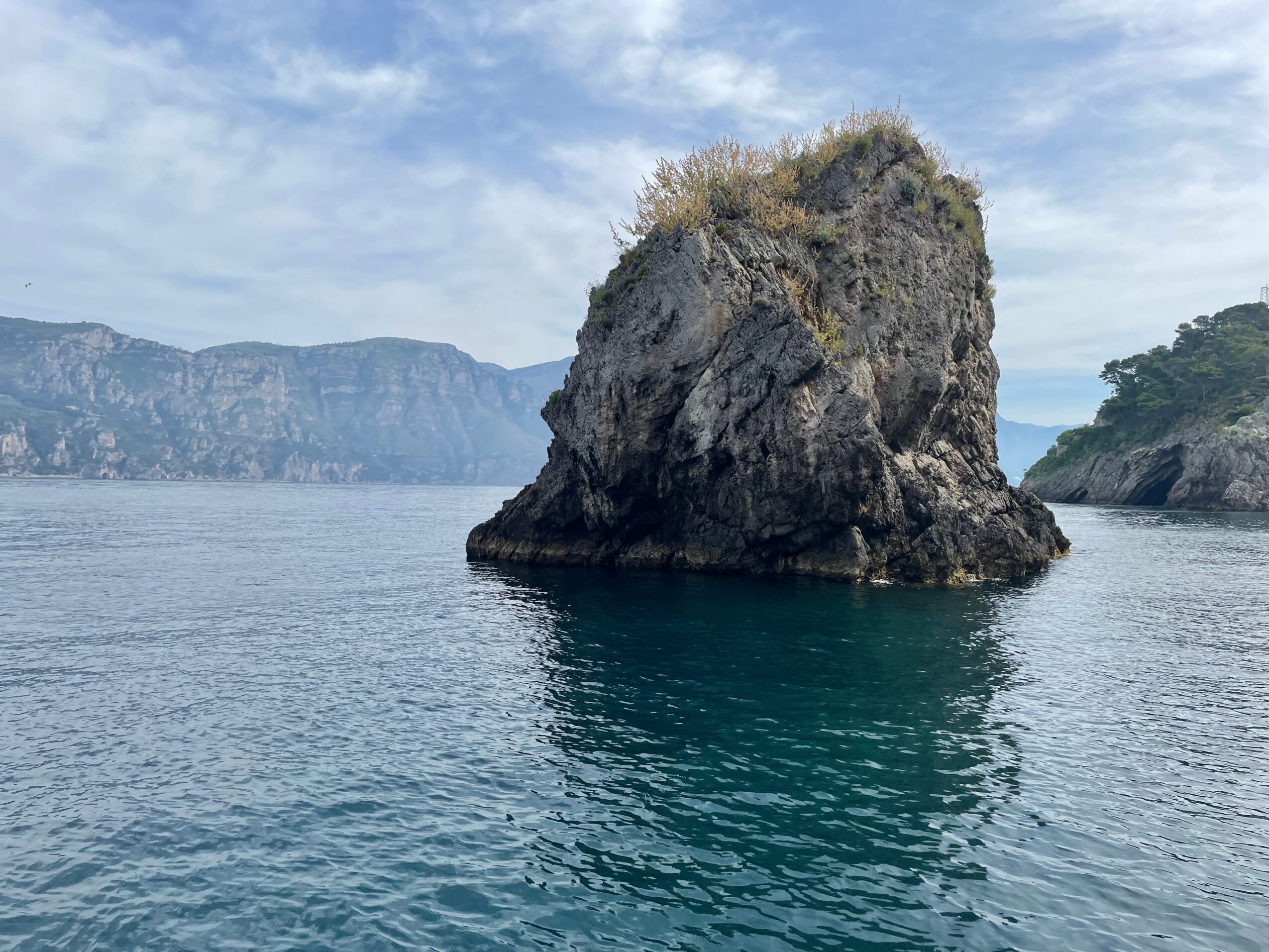 Large rock formation in water with distant cliffs and partly cloudy sky, calm sea surface, and some vegetation on top.