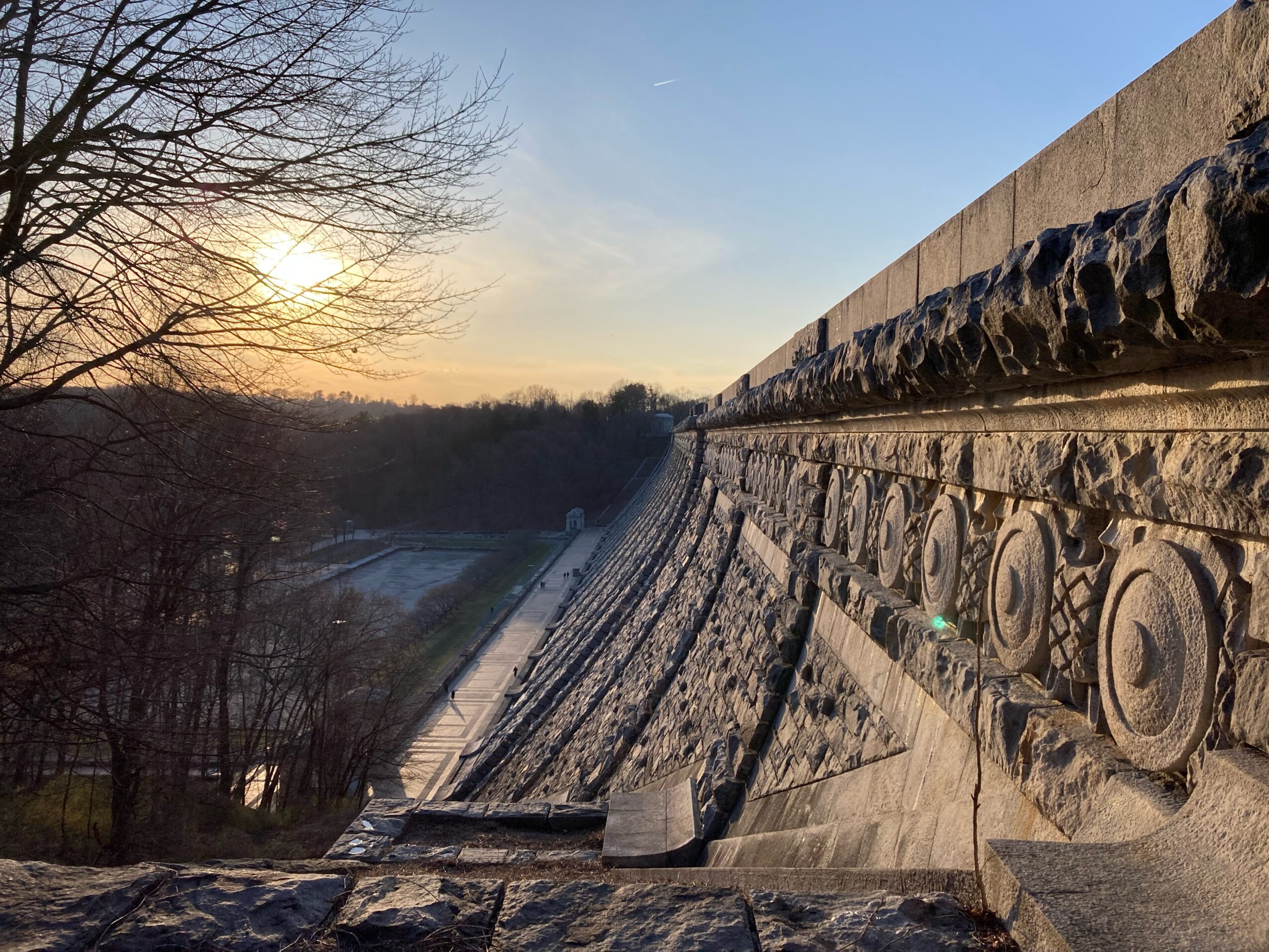 Sunset over a landscape with a stone wall and trees, with a river or road below, viewed from an elevated position.