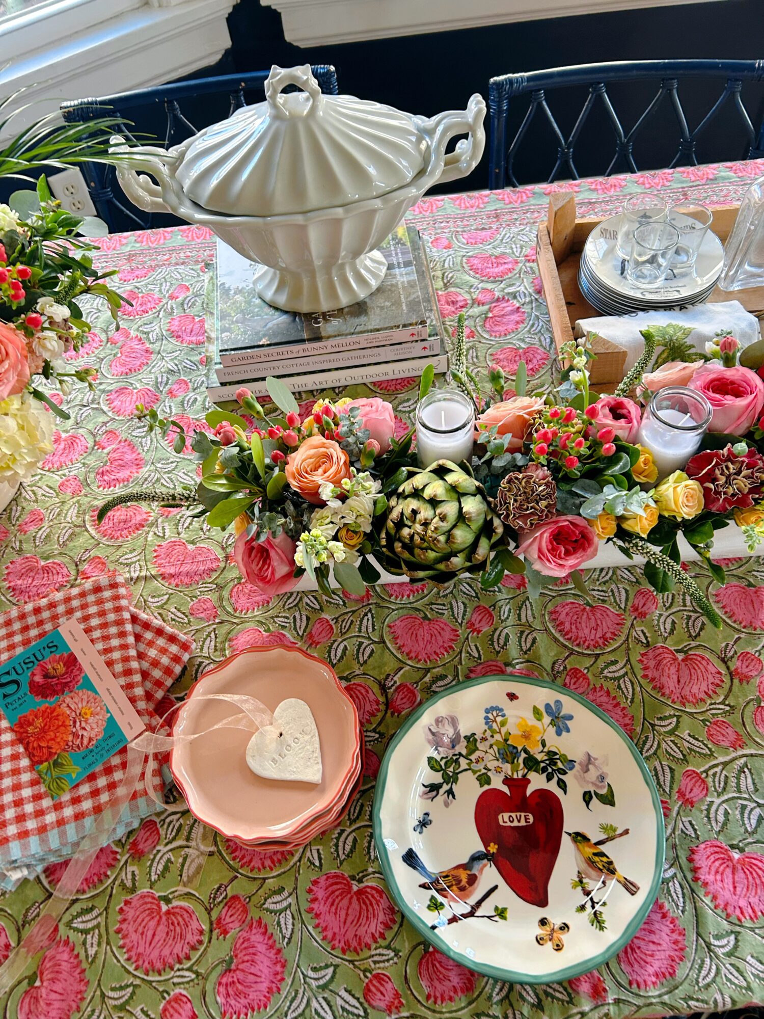 Table with floral tablecloth, decorative centerpiece, teapot, and plates, set for a meal or celebration, with pink and green colors.