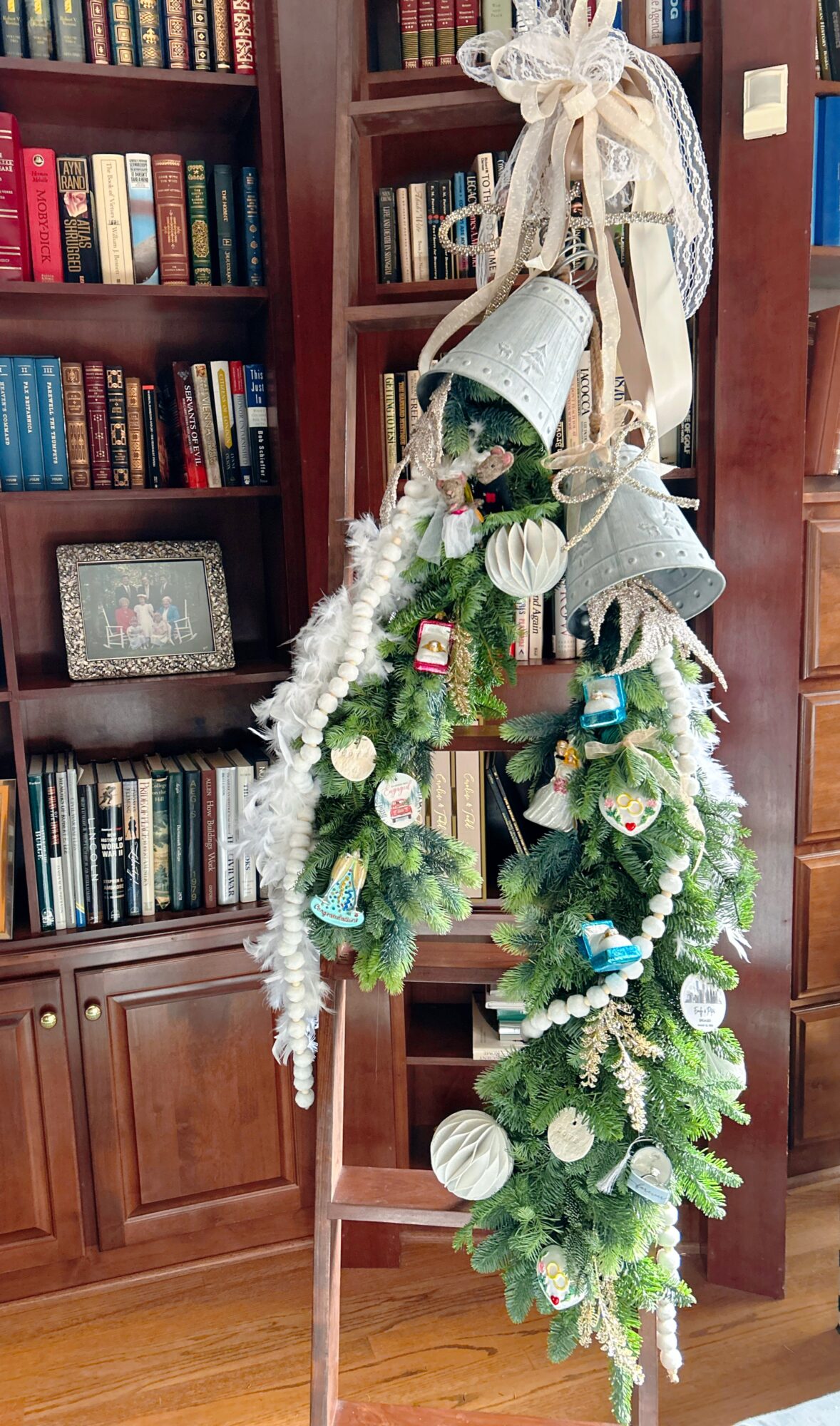 Decorated Christmas wreath hanging from a wooden ladder, with ornaments, ribbons, and greenery, in a room with bookshelves.