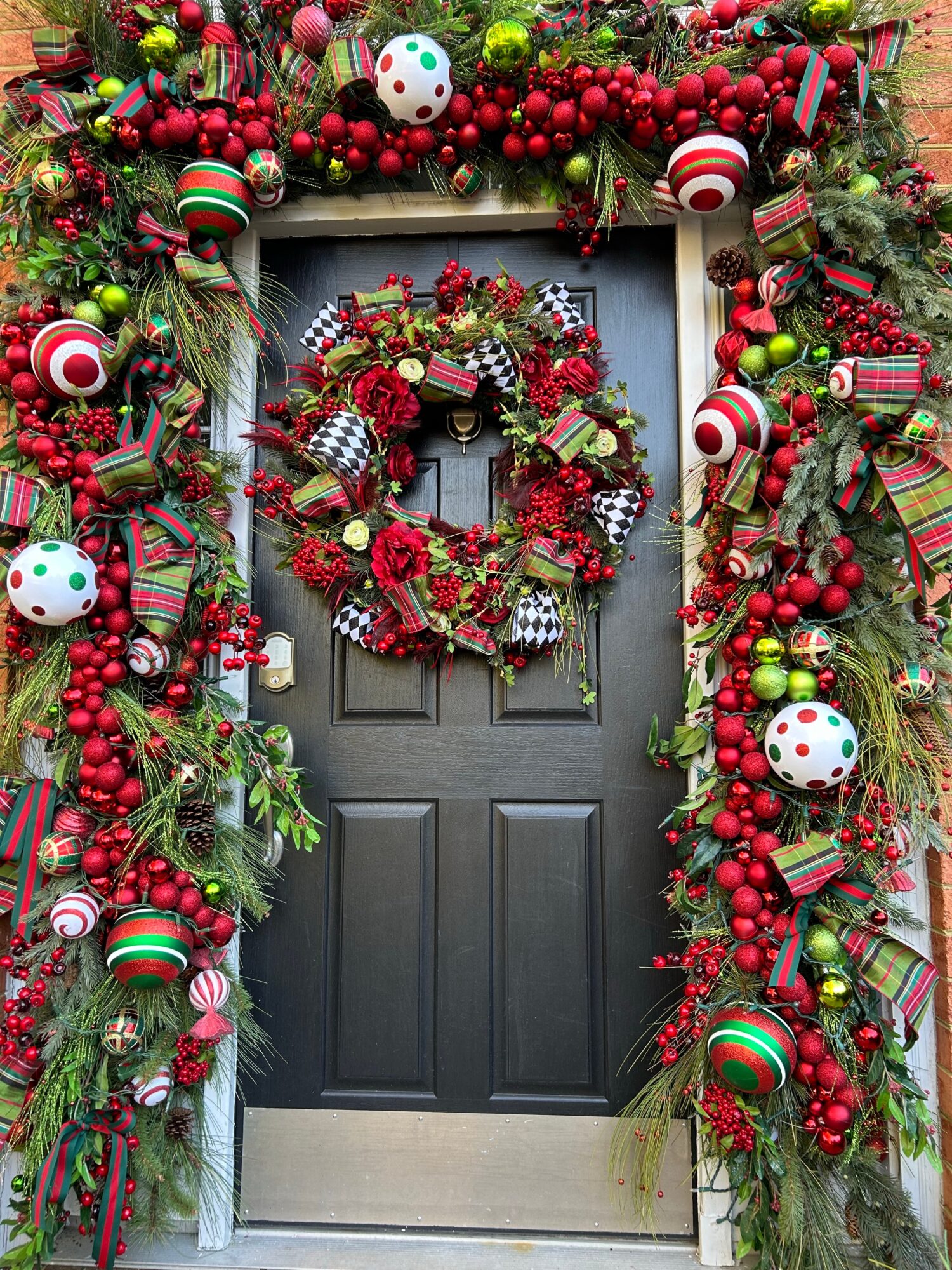 Decorated front door with a Christmas wreath and festive garland with ornaments and ribbons, surrounded by greenery.
