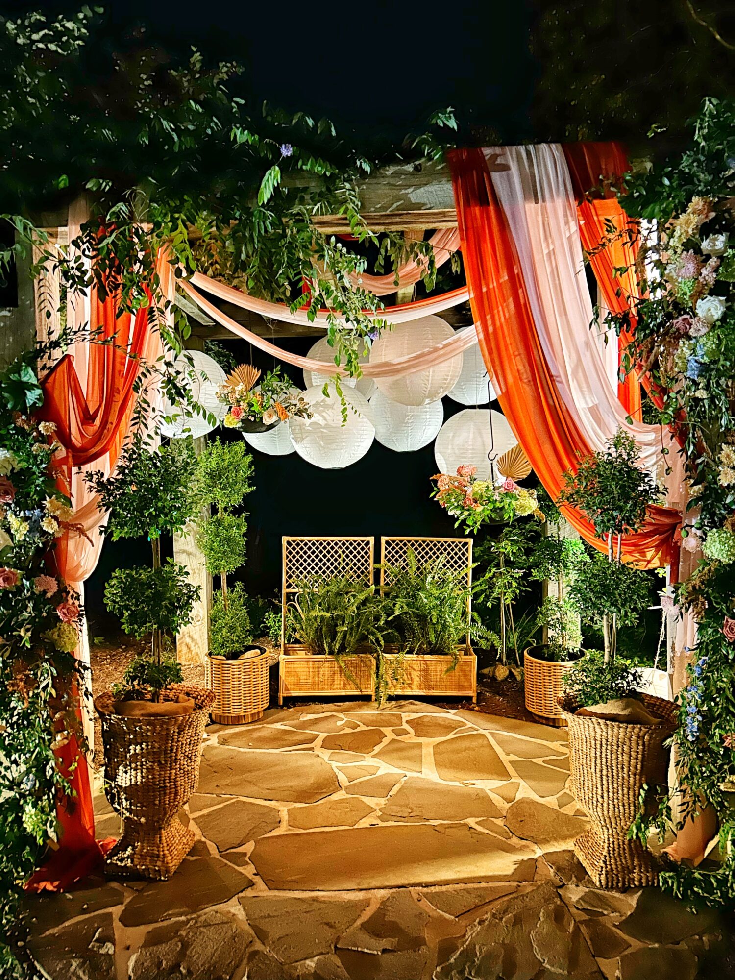 Decorated outdoor space with draped orange and white fabric, hanging lanterns, potted plants, and a stone pathway.