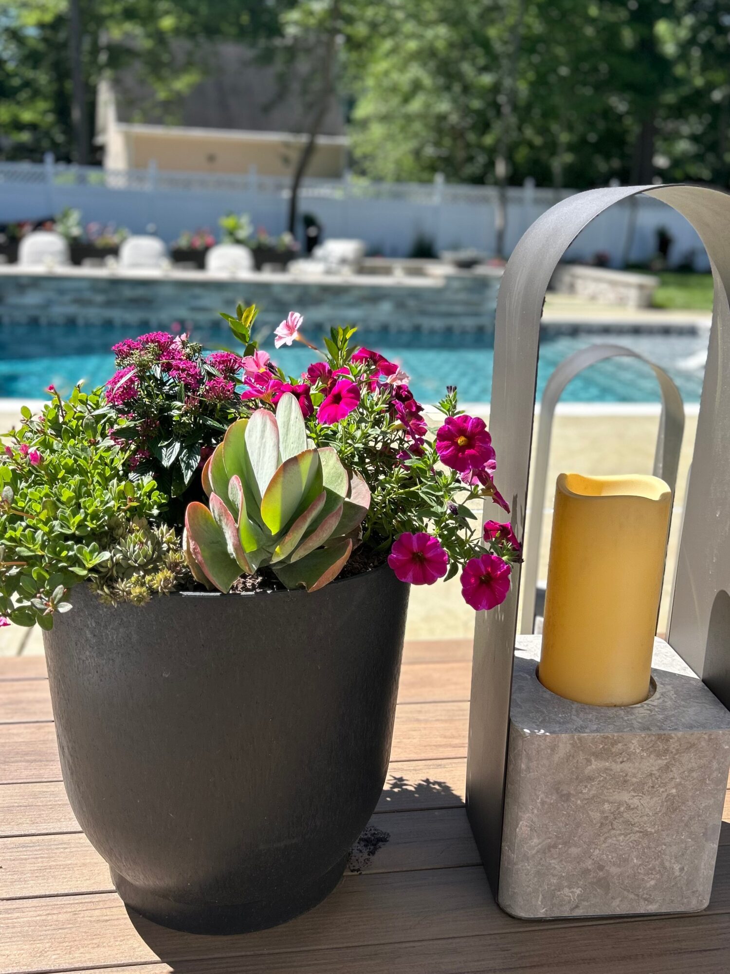 Flower pot with pink, white, and green plants next to a yellow candle on a wooden deck near a pool.