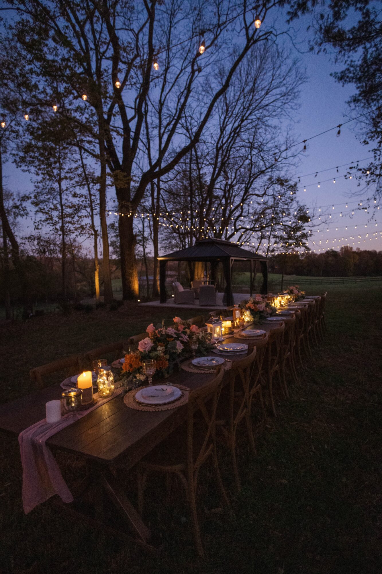 Long outdoor dining table with candles and flowers, trees and a pavilion in the background during evening, twilight sky.