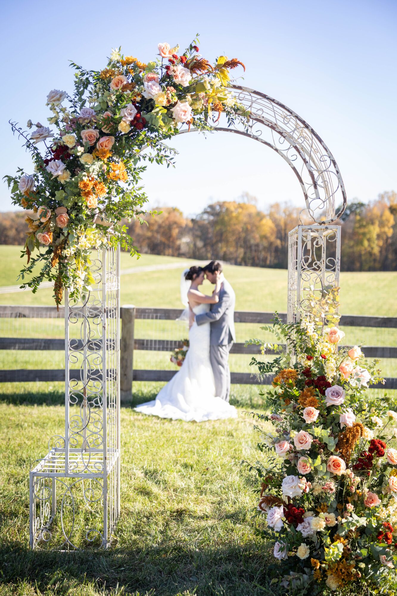 Couple kissing under a decorated arch outdoors with flowers, fence, and trees in the background.